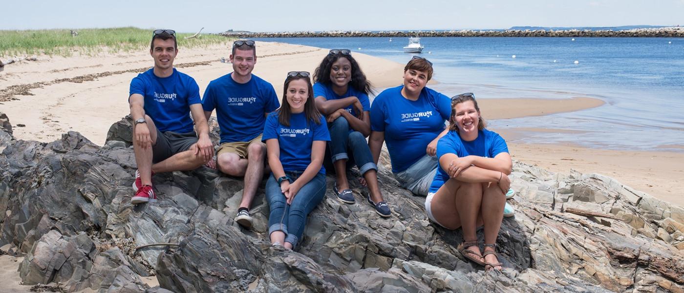 A group of U N E student tour guides wear matching blue shirts while sitting on a rocky beach